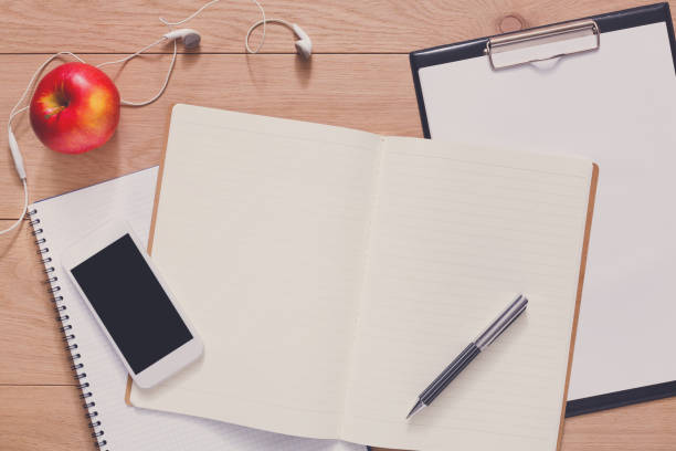 Notepad, diary, mobile phone and apple fruit. Office or student's supplies on modern wooden desk. Working table top view. Education or job background with copy space on paper sheet