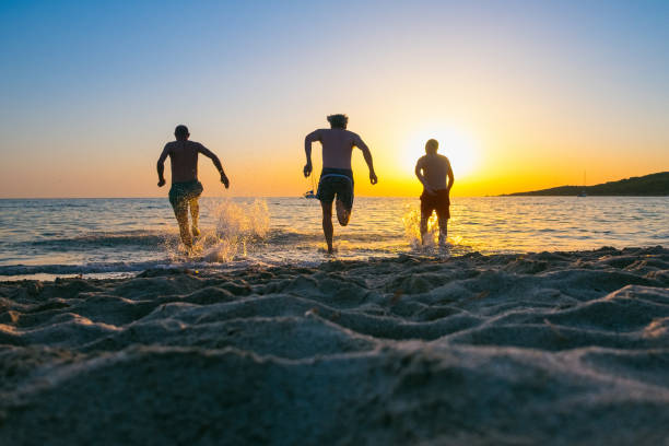 grupo de amigos correndo para o mar ao pôr do sol - silhouette three people beach horizon - fotografias e filmes do acervo