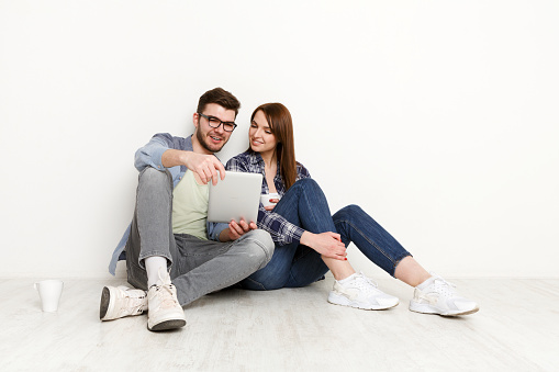 Wasting money for shopping. Couple at home sitting on floor and using digital gadget and drinking coffee together, white background