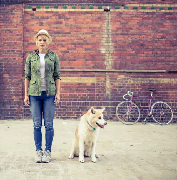 Hipster young beautiful girl with vintage road bike in city relaxing with akita inu dog, urban scene. Woman commuter cycling on fixed gear bike in town, retro city street industrial background.