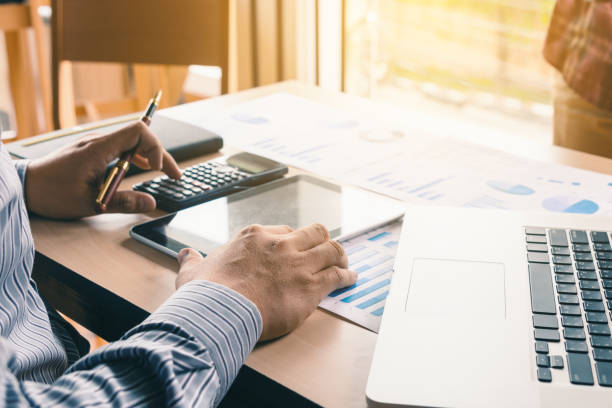 Asian businessman pressing button on calculator and calculate about analyze with looking data on tablet screen. stock photo