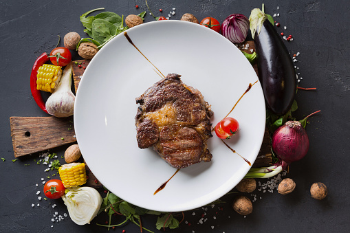 Rib eye steak on white plate on wooden desk with vegetables and spices border on dark background, top view.