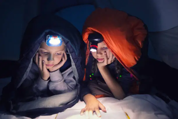 Two children in a tent on camping wrapped in a sleeping bag reading a book with flashlight on their head at night