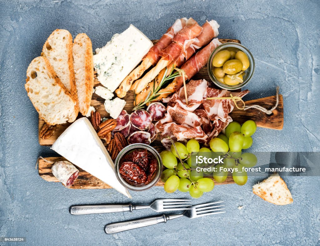 Cheese and meat appetizer selection or wine snack set. Variety of  , salami, prosciutto, bread sticks, baguette, honey, grapes, olives, sun-dried tomatoes, pecan nuts over grey concrete textured backdrop, top vie Cheese and meat appetizer selection. Prosciutto di Parma, salami, bread sticks, baguette slices, olives, sun-dried tomatoes, grapes and nuts on rustic wooden board over grey concrete textured backdrop, top view Cheese Stock Photo
