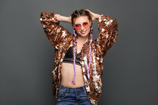 Young stylish woman with braids wearing golden jacket and sunglasses posing on gray background