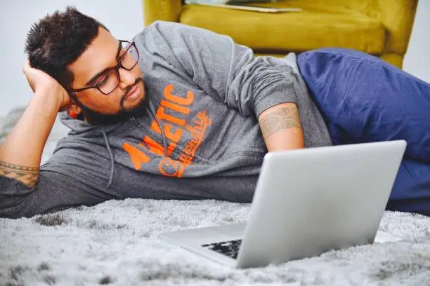 A Handsome tattooed man lying near a chair, working on his laptop computer.