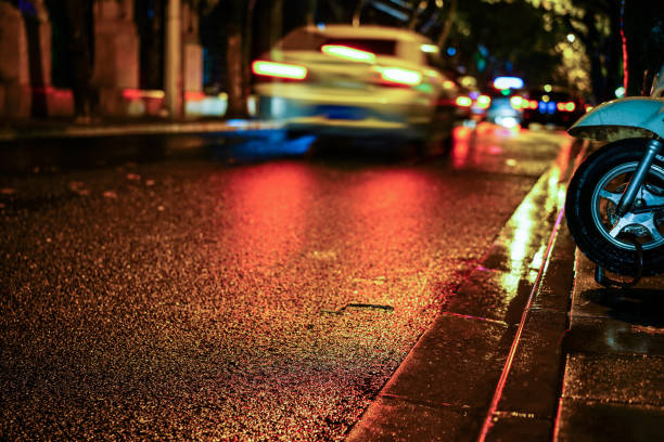 Rainy night in the big city, light from the shop windows reflected on the road on which cars travel. View from the level of asphalt.