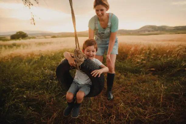 Photo of young mother bonding with her son, swinging him on a tire swing they have built together. They enjoy beautiful summer day, outdoors in the nature.