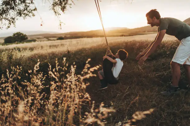 Photo of young father bonding with his son, swinging him on a tire swing they have built together. They enjoy beautiful summer day, outdoors in the nature.