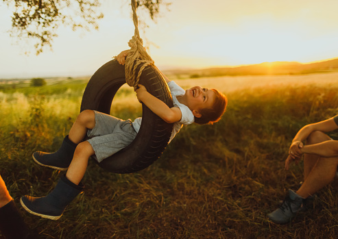 Photo of young family with little boy, who is swinging on a tire swing as they spend time together, outdoors in the nature