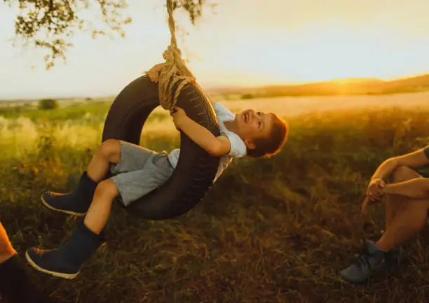 Photo of young family with little boy, who is swinging on a tire swing as they spend time together, outdoors in the nature