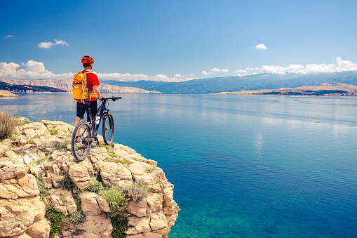 Mountain biker looking at view and traveling on bike in summer sea landscape. Man rider cycling MTB on rocky yellow trail. Fitness motivation, inspiration in beautiful inspirational view.