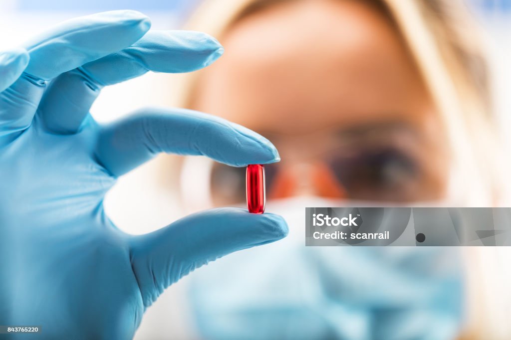 Young attractive female scientist holding a red transparent pill Young attractive female scientist with protective eyeglasses and mask holding a red transparent pill with fingers in gloves in the pharmaceutical research laboratory Medicine Stock Photo
