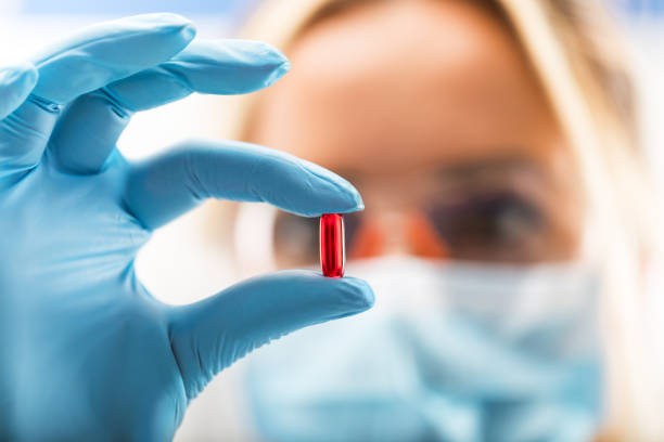 Young attractive female scientist holding a red transparent pill Young attractive female scientist with protective eyeglasses and mask holding a red transparent pill with fingers in gloves in the pharmaceutical research laboratory pharmaceutical compounding stock pictures, royalty-free photos & images