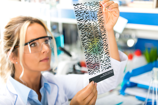 Young attractive female scientist with protective eyeglasses examining DNA autoradiogram test results in the scientific biochemical laboratory