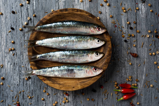 raw sardines on a rustic wooden table high-angle shot of some raw sardines in a wooden plate, placed on a gray rustic wooden table sprinkled with peppercorns sardine photos stock pictures, royalty-free photos & images