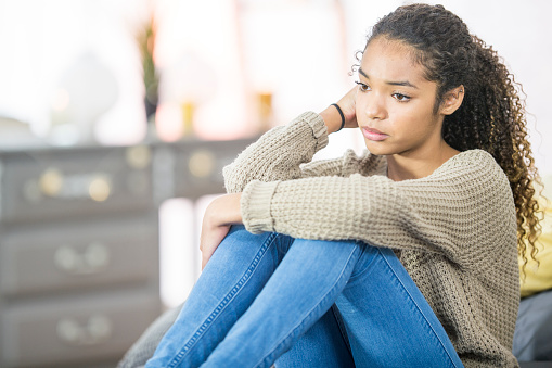 A teenage girl of African descent is indoors in a bedroom. She is wearing casual clothing. She is sitting on her bed and looking sad.