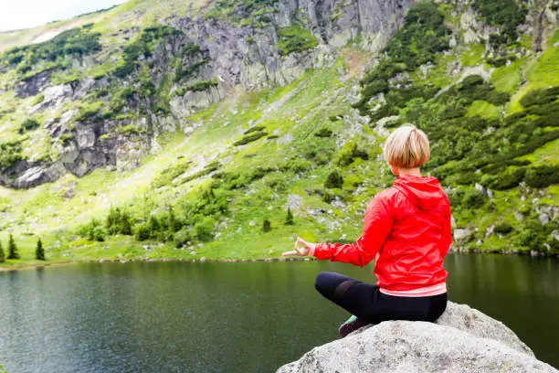 Woman meditating in yoga pose in front of mountain lake. Female hiker relaxing in beautiful inspirational mountains landscape. Healthy lifestyle outdoors in nature concept.