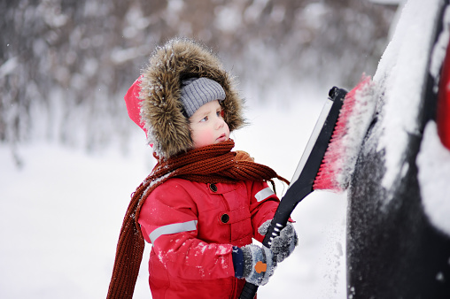 Cute little child helping to brush a snow from a car. Toddler boy holding tool for cleaning