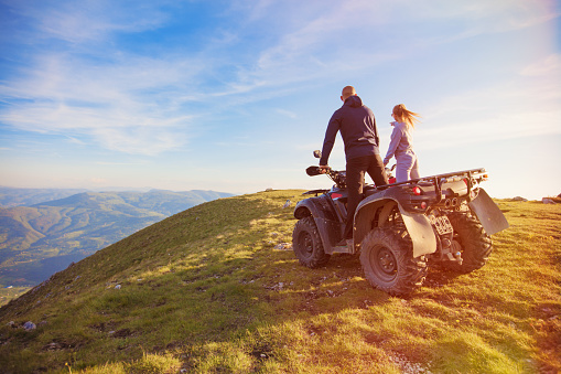 Rear view of young pair near atv. Man is showing something in distance to her girlfriend. Blurred autumn landscape mighty mountains and forests on background
