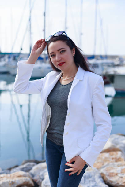 Portrait of woman dressed in nautical style in yacht marina Vertical portrait of woman dressed in nautical style and wearing white jacket and string of pearls  with her hand touching eye glasses on her head and yacht marina blurred in background dispelled stock pictures, royalty-free photos & images