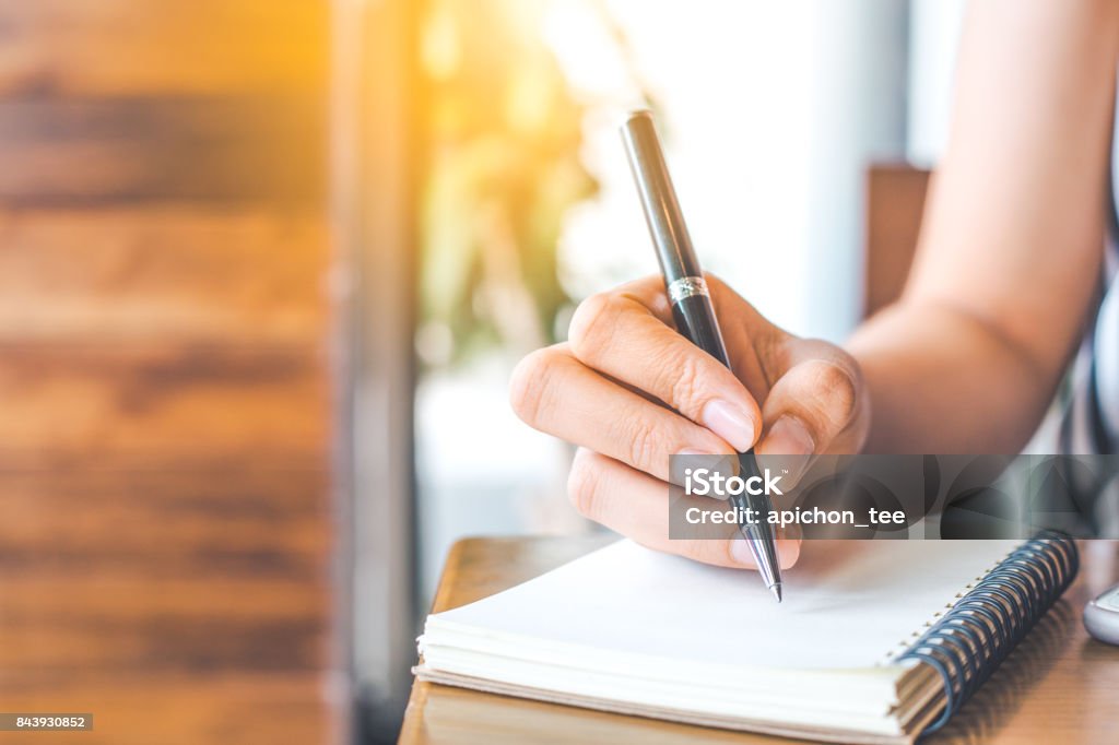 woman's hand is writing on a blank notepad with a pen on a wooden desk. List Stock Photo