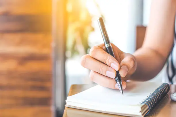 Photo of woman's hand is writing on a blank notepad with a pen on a wooden desk.