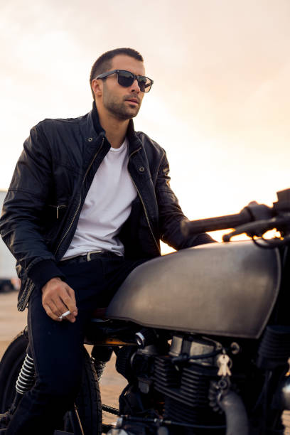 Brutal man sit on cafe racer custom motorbike. Beautiful happy rider man with beard and mustache in black biker jacket, jeans and fashion sunglasses smoking cigaret sit on classic style cafe racer motorbike at sunset. Brutal fun urban lifestyle. cafe racer stock pictures, royalty-free photos & images