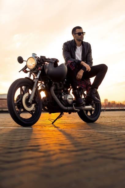 Brutal man sit on cafe racer custom motorbike. Handsome rider man with beard and mustache in black leather biker jacket and sunglasses sit on classic style cafe racer motorcycle. Bike custom made in vintage garage. Brutal fun urban lifestyle. cafe racer stock pictures, royalty-free photos & images