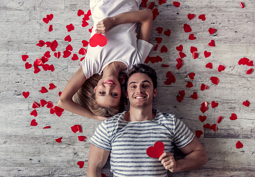Top view of beautiful young couple holding red paper hearts, looking at camera and smiling while lying on the floor