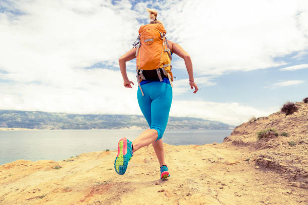 Woman running with backpack on rocky trail at seaside and mountains. Young woman training and trail running on mountain rocky dirt path looking at beautiful inspirational landscape view.