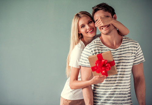 Beautiful young woman is holding a gift box and covering her boyfriend eyes making a surprise, both are smiling, on gray background
