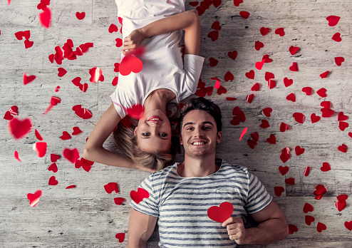 Top view of beautiful young couple holding red paper hearts, looking at camera and smiling while lying on the floor