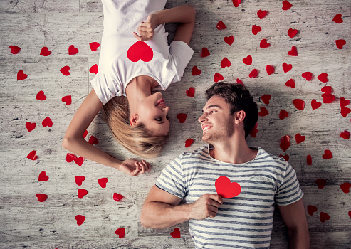 Top view of beautiful young couple holding red paper hearts, looking at each other and smiling while lying on the floor