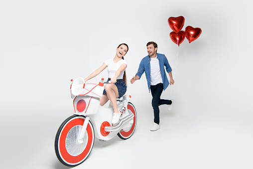 girlfriend riding handmade electric bike while her boyfriend running for her with balloons, isolated on grey