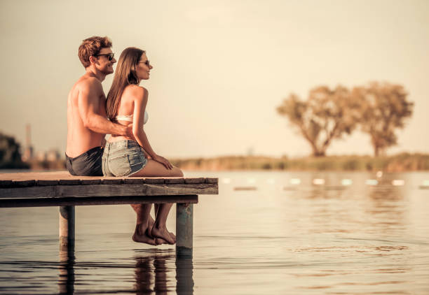 Happy couple is hugging, enjoying the seaside view and smiling while sitting on pier at the sea