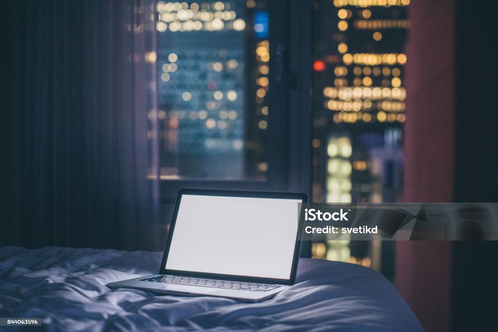 Always online Laptop on bed in dark hotel room. Laptop Stock Photo