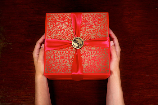 Hands presenting a gift box with red ribbon on dark wooden background.