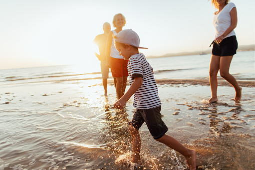 Photo of a little boy spending summer days by the ocean surrounded by his family; he is in a foreground, while the rest of the people are in the back, defocused