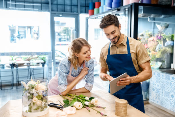 florists with digital tablet in flower shop smiling young florists in aprons using digital tablet while working together in flower shop florist stock pictures, royalty-free photos & images