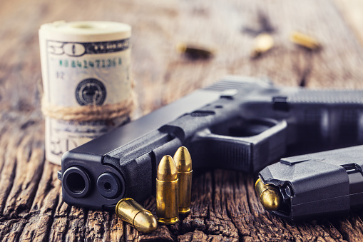 Gun and money. 9 mm pistol gun bullets strewn and roll dollar banknotes on rustic oak table.