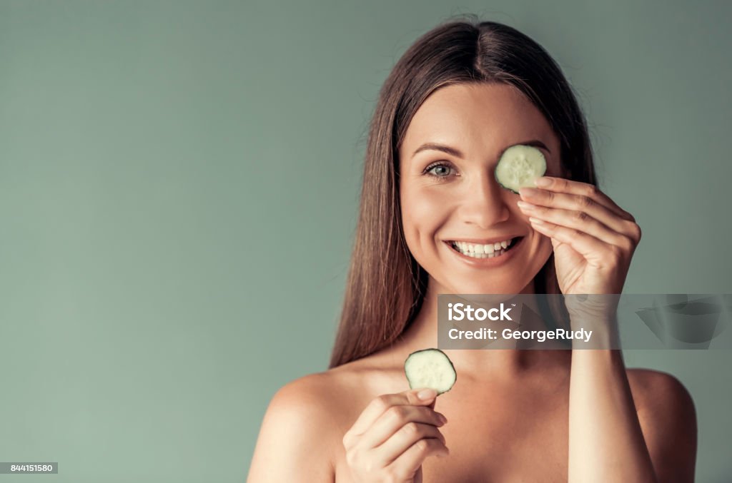 Young woman, beauty Beautiful woman with bare shoulders is holding slices of cucumber, looking at camera and smiling, on gray background Cucumber Stock Photo