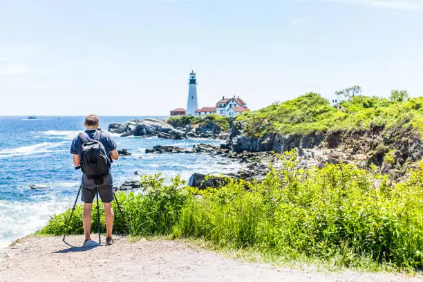Back of man photographer taking pictures on tripod of Portland Head Lighthouse in Fort Williams park in Maine during summer day