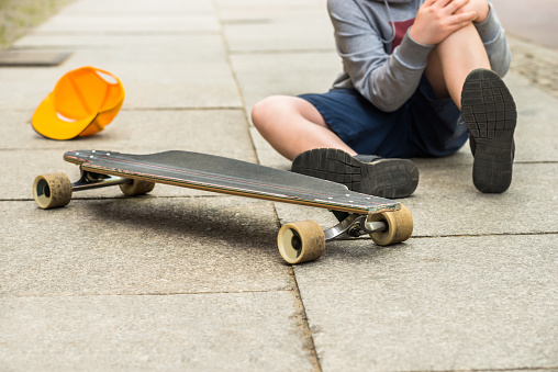 Boy With Knee Injury Sitting Near Skateboard On Sidewalk