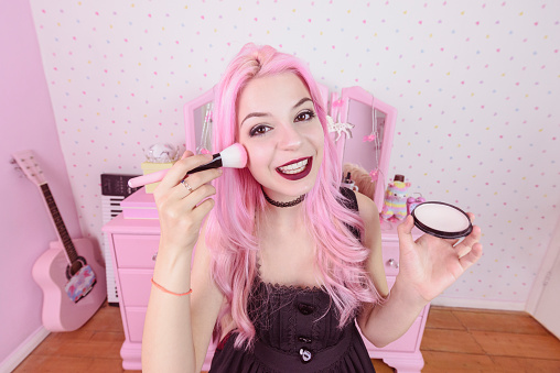 Woman with dyed hair in bedroom holding make up brush and putting on cosmetics, smiling with cheerful expression
