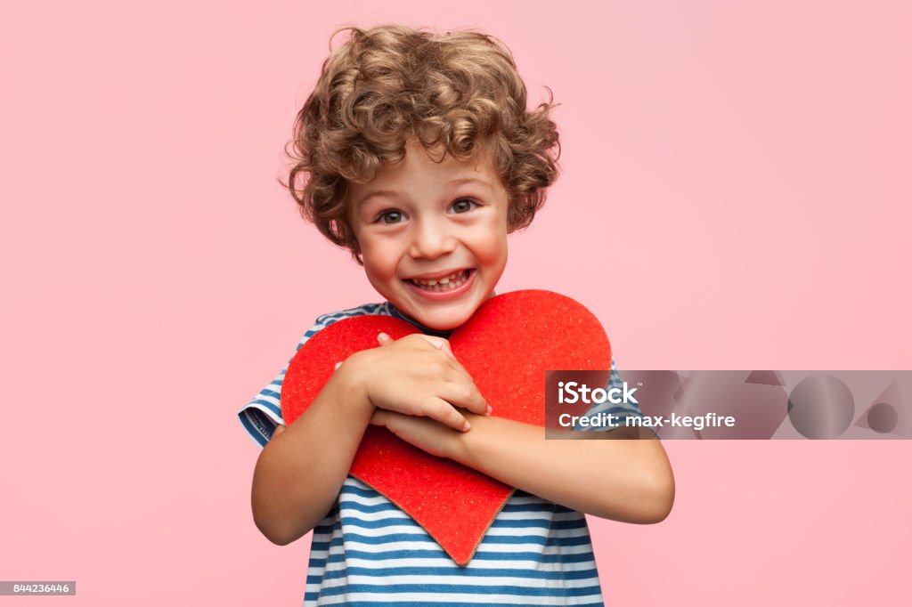 Charming boy posing with heart Little curly boy holding heart application and laughing at camera on pink background. Child Stock Photo