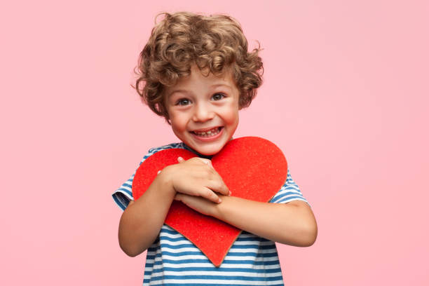 Charming boy posing with heart Little curly boy holding heart application and laughing at camera on pink background. charming photos stock pictures, royalty-free photos & images
