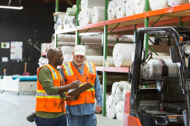 Two men working in carpet warehouse, with forklift Two multi-ethnic men working in a carpet warehouse wearing safety vests. The African-American man is holding a clipboard and talking. They are standing next to a forklift. carpet factory photos stock pictures, royalty-free photos & images