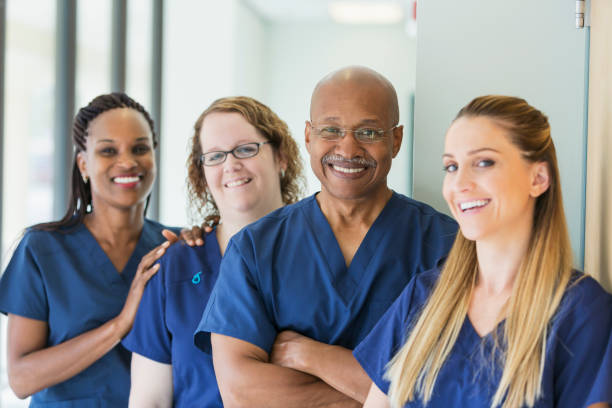 Man leading team of multi-ethnic medical professionals A team of four multi-ethnic medical professionals standing in a corridor, wearing blue scrubs, smiling at the camera. The focus is on the African-American senior man, in his 60s. female nurse photos stock pictures, royalty-free photos & images