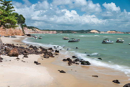 Long exposure of Pipa Beach (Praia da Pipa) with multiple tourist boats floating on a sunny day with cloudy moody sky. Sandy beach with cliffs and dunes on the background.